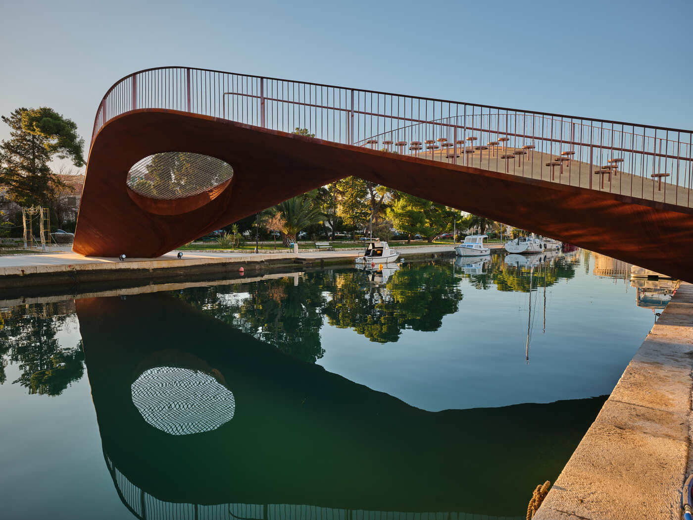 Pješački most na Foši u Trogiru – čelična ljuskasta konstrukcija s otvorom i mrežom, odraz u mirnoj vodi.